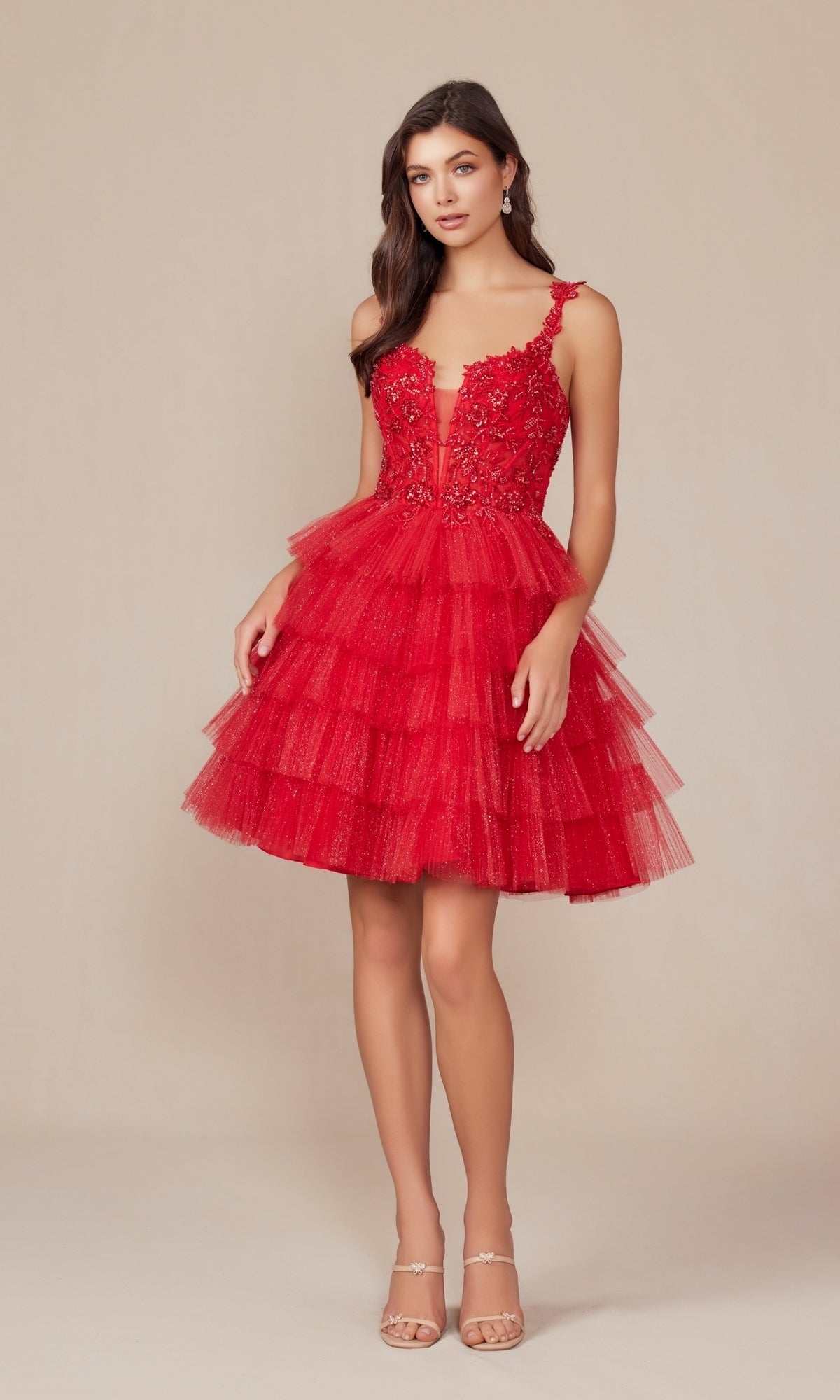 Woman wearing the Glitter Babydoll Homecoming Dress: Nox Anabel E904—short, red, sleeveless with a floral applique bodice and tiered skirt—standing against a plain beige background.